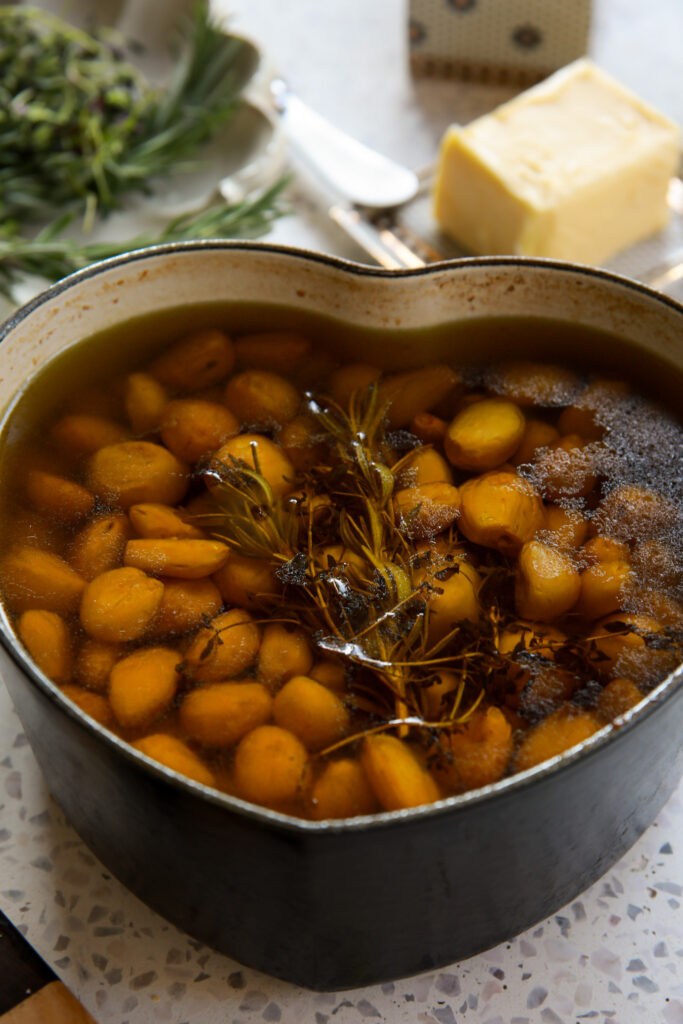 Garlic confit recipe cooking in a heart-shaped pot filled with olive oil, herbs, and caramelized cloves.