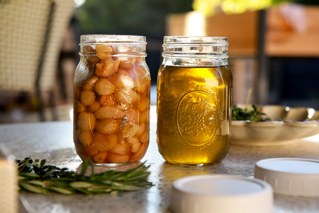 Two mason jars filled with garlic confit and olive oil, styled with rosemary sprigs on a white counter in warm light.