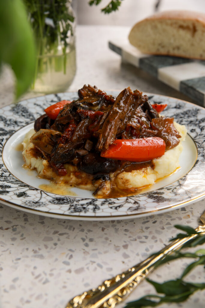 Slow cooker creamy mushroom pot roast served over mashed potatoes with carrots on a patterned plate.